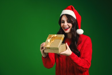 Photo of Attractive woman in Santa hat opening Christmas gift on green background, space for text