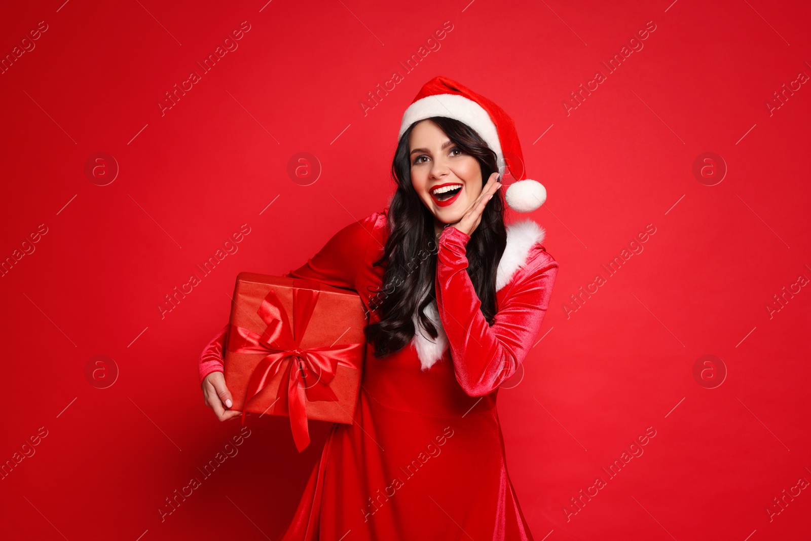 Photo of Beautiful woman in Mrs Claus costume with Christmas gift on red background