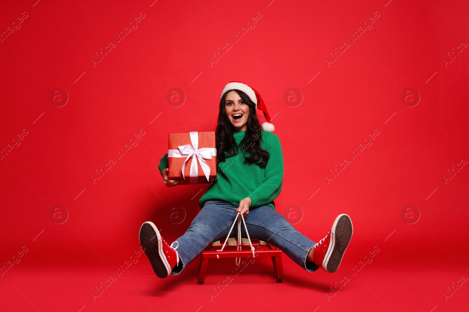 Photo of Happy woman in Santa hat with Christmas gift on sledge against red background