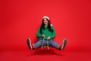 Photo of Happy woman in Santa hat on sledge against red background. Christmas celebration