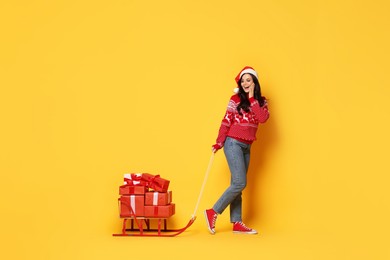 Woman in Santa hat with Christmas gifts on sledge against yellow background