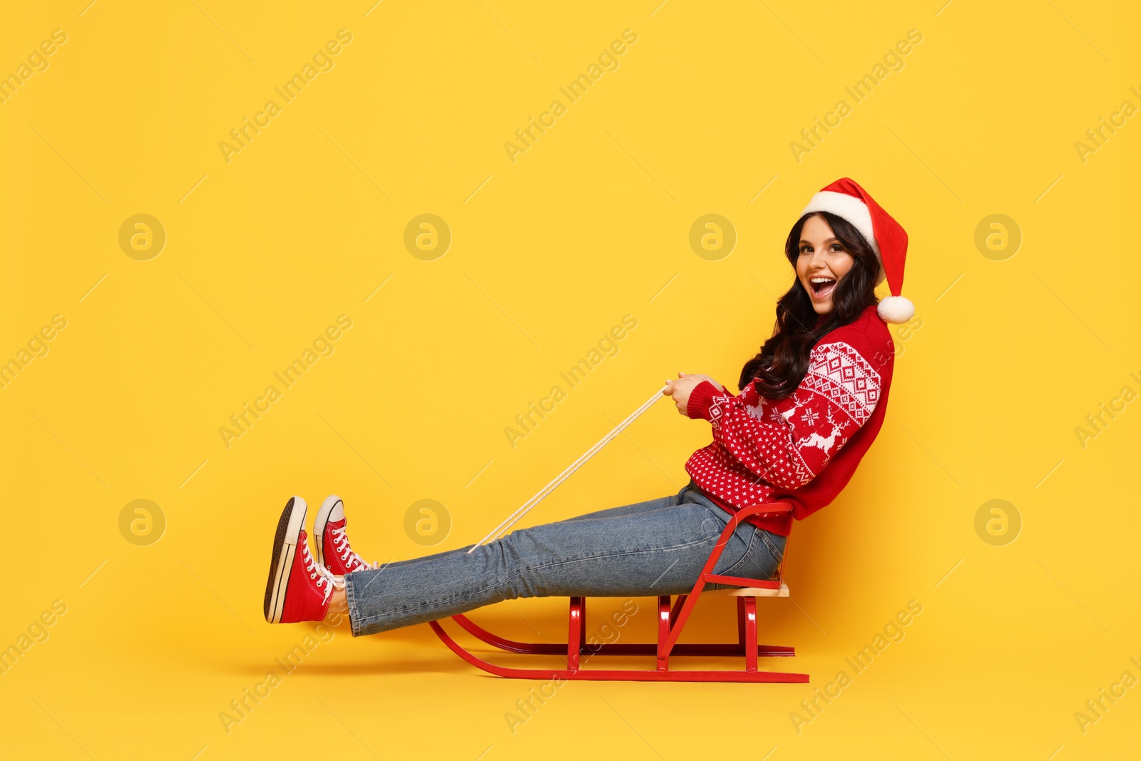 Photo of Woman in Santa hat on sledge against yellow background, space for text. Christmas celebration