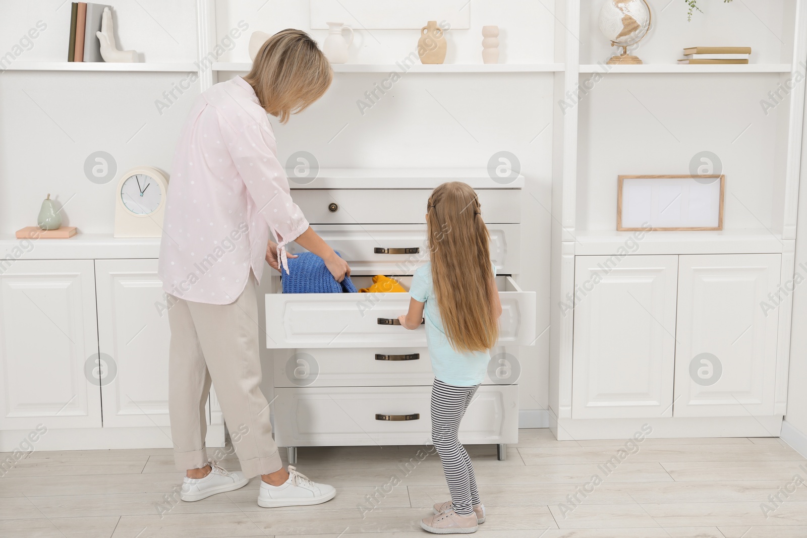 Photo of Little girl helping her mother putting clothes into drawers at home