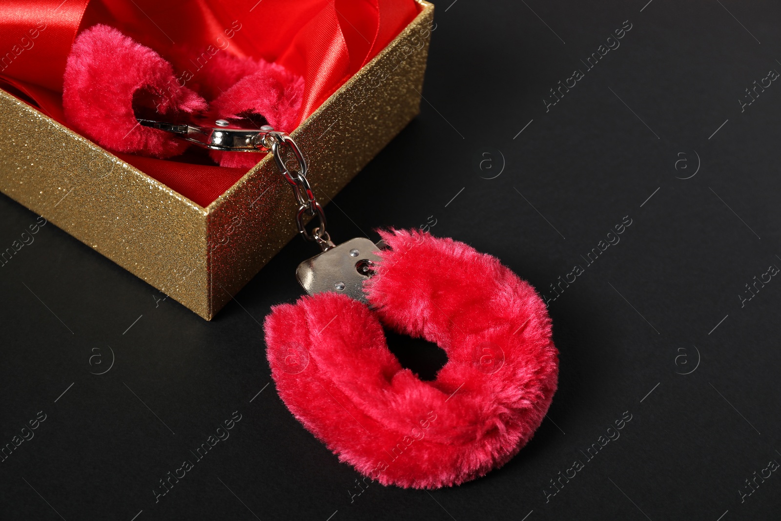 Photo of Bright fluffy handcuffs and gift box on black background, space for text