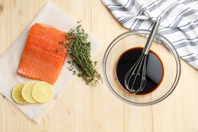 Photo of Soy sauce in bowl, whisk, salmon fillet, thyme and lemon slices on wooden table, flat lay