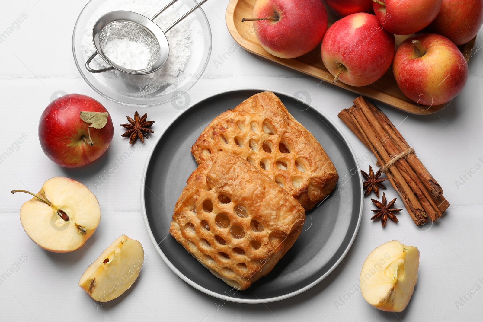 Photo of Delicious puff pastries, apples, cinnamon, anise stars and powdered sugar on white tiled table, top view