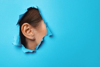 Woman showing her ear through hole in light blue paper, closeup. Space for text