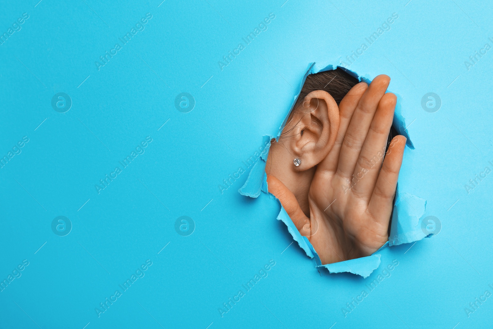 Photo of Woman showing hand to ear gesture through hole in light blue paper, closeup. Space for text