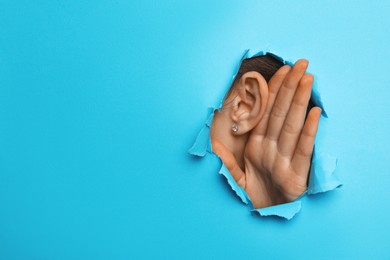 Photo of Woman showing hand to ear gesture through hole in light blue paper, closeup. Space for text