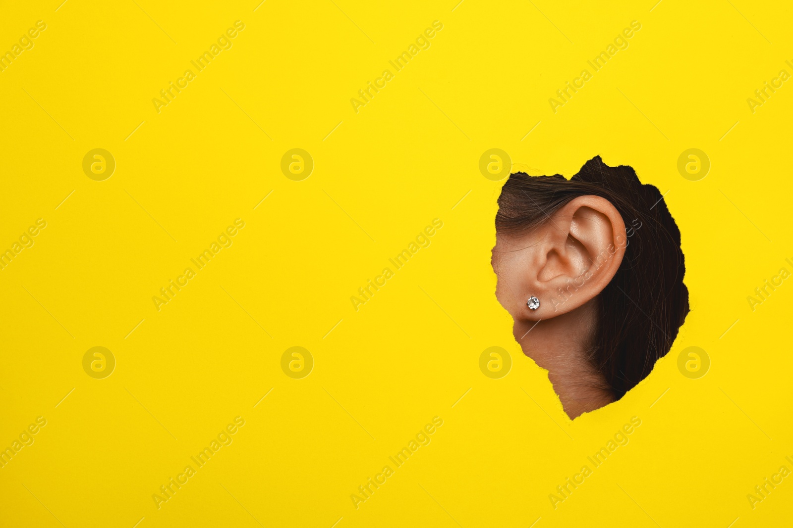 Photo of Woman showing her ear through hole in yellow paper, closeup. Space for text