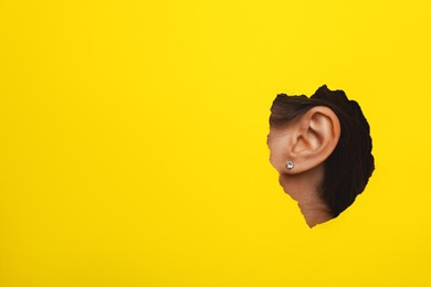 Woman showing her ear through hole in yellow paper, closeup. Space for text