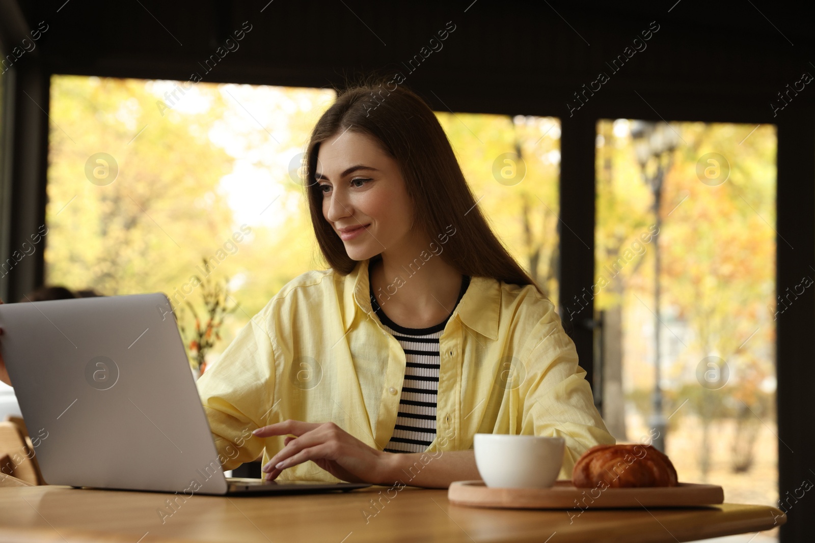 Photo of Woman with coffee and croissant working on laptop at table in cafe