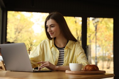 Woman with coffee and croissant working on laptop at table in cafe