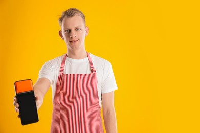 Happy young man in apron with payment terminal on yellow background