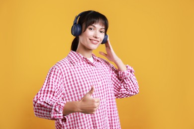 Photo of Happy woman in headphones showing thumbs up on orange background, space for text. Like gesture