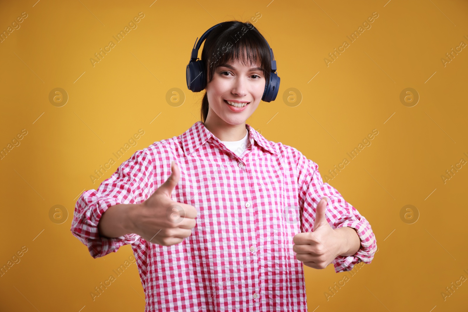 Photo of Happy woman in headphones showing thumbs up on orange background. Like gesture