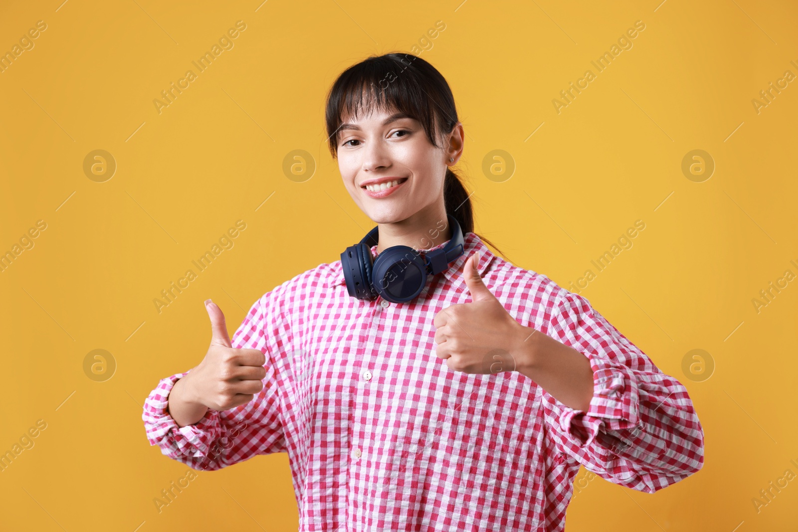 Photo of Happy woman with headphones showing thumbs up on orange background. Like gesture