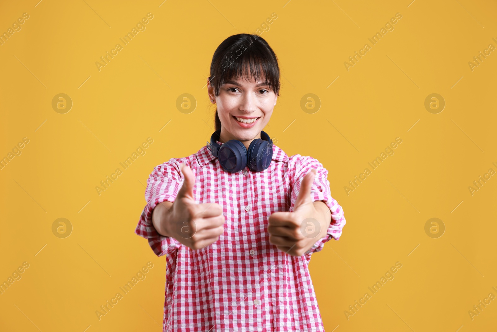 Photo of Happy woman with headphones showing thumbs up on orange background. Like gesture