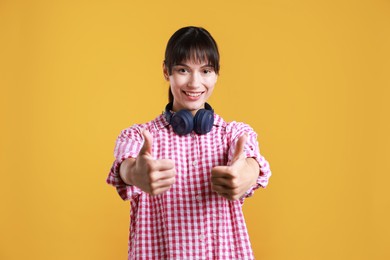 Photo of Happy woman with headphones showing thumbs up on orange background. Like gesture