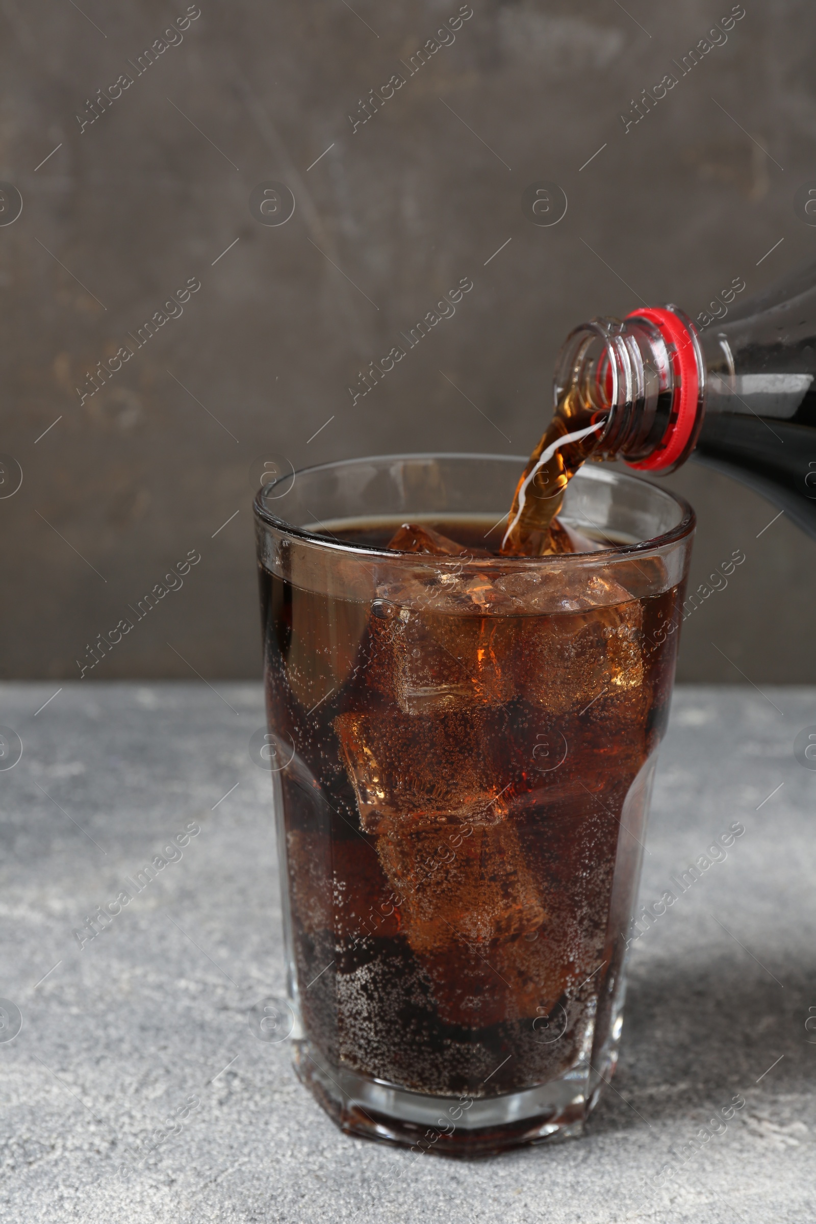 Photo of Pouring refreshing cola from bottle into glass on light textured table against grey background, closeup