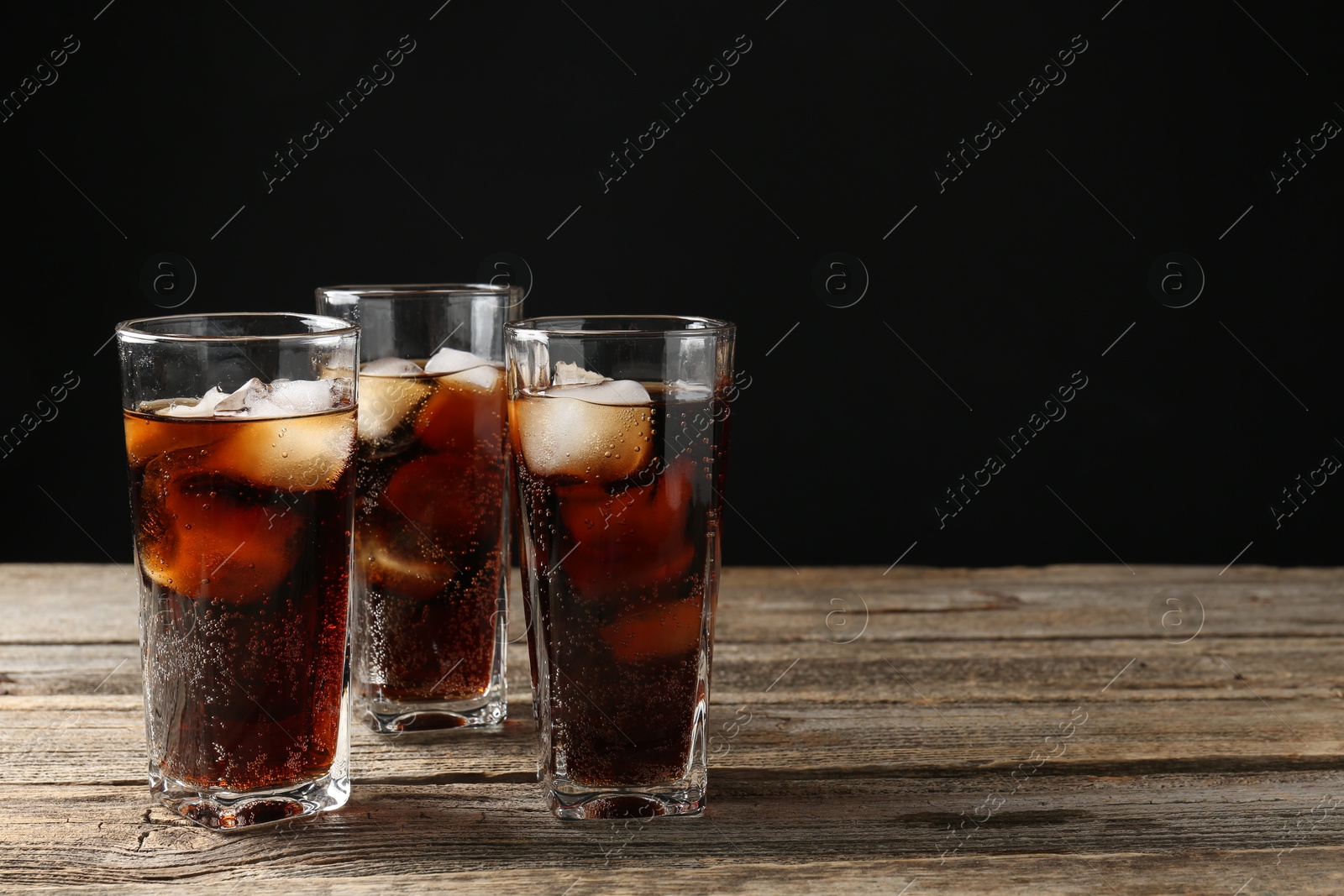 Photo of Refreshing cola with ice cubes in glasses on wooden table against black background, closeup. Space for text