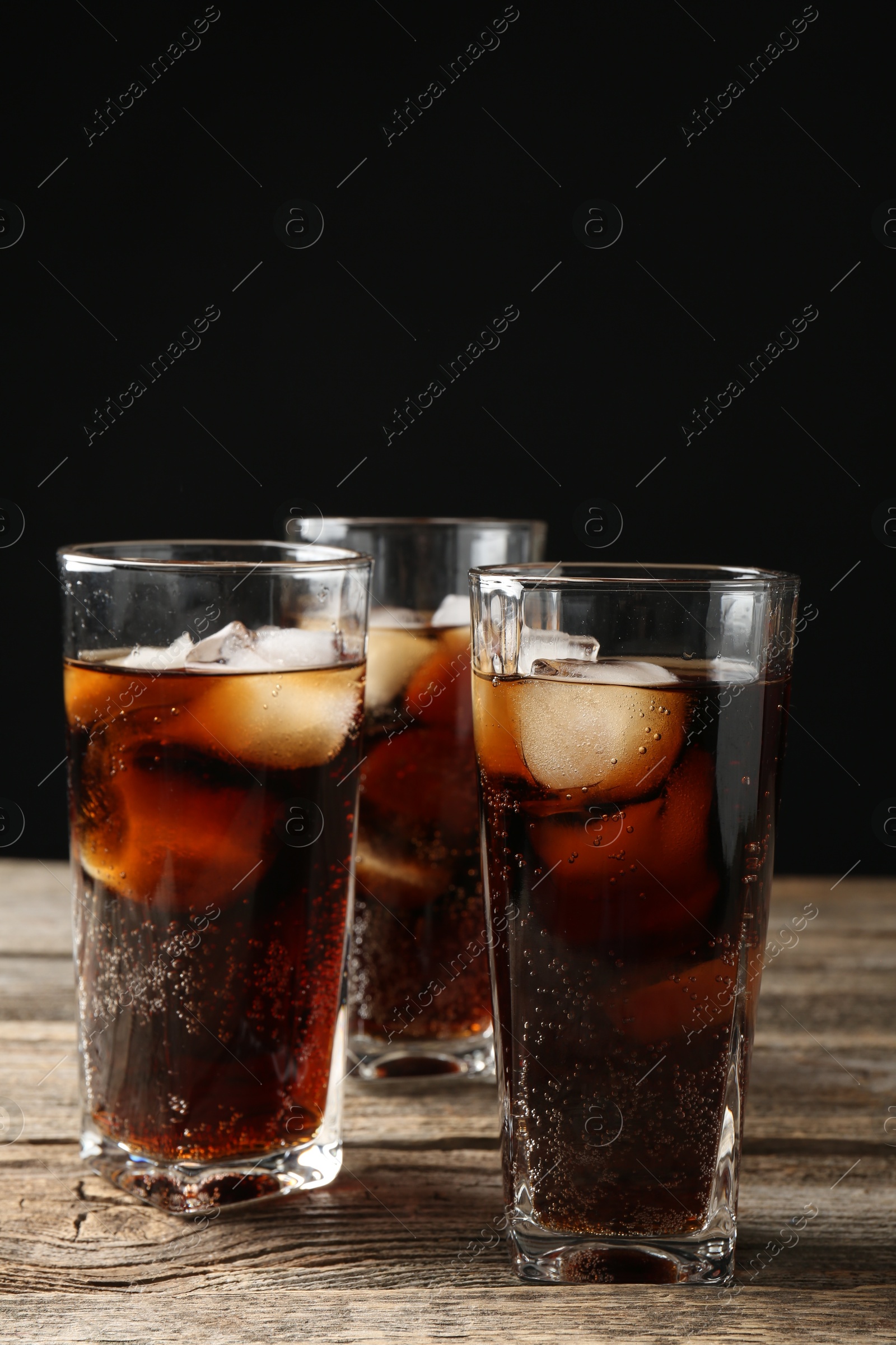 Photo of Refreshing cola with ice cubes in glasses on wooden table against black background, closeup