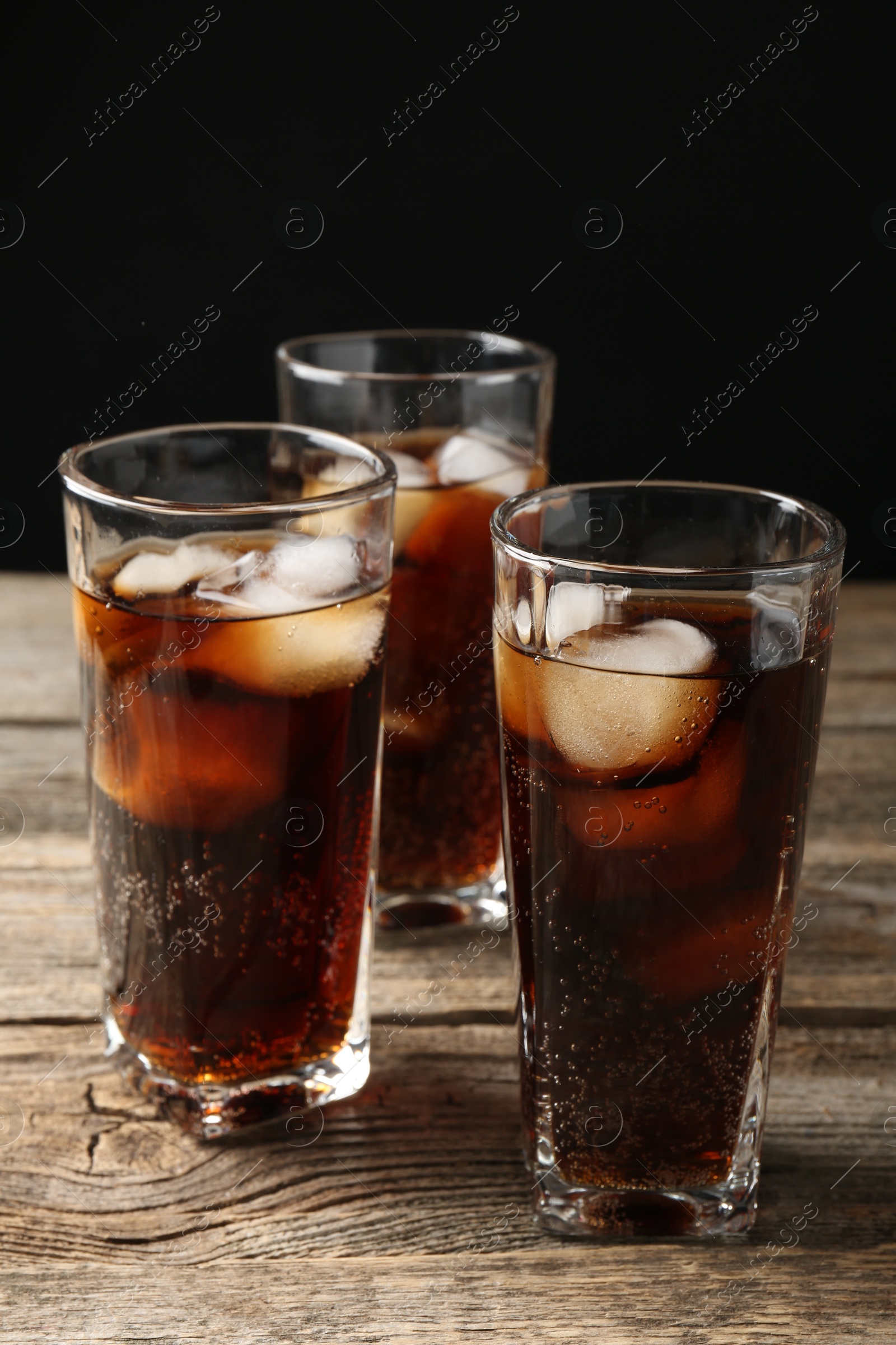 Photo of Refreshing cola with ice cubes in glasses on wooden table against black background, closeup