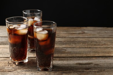 Photo of Refreshing cola with ice cubes in glasses on wooden table against black background, closeup. Space for text