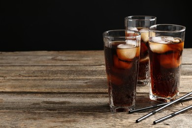 Photo of Refreshing cola with ice cubes in glasses and drinking straws on wooden table against black background. Space for text