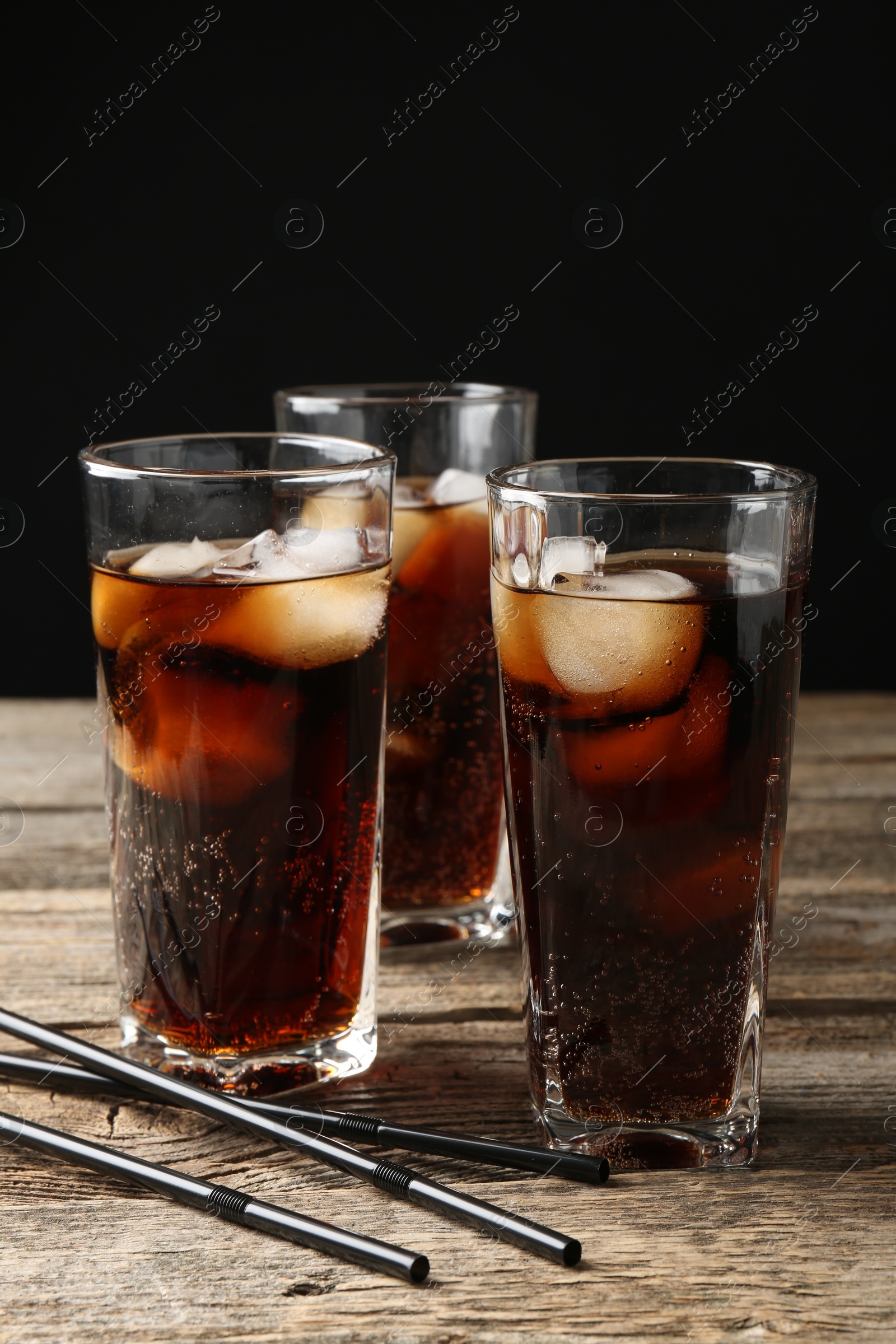 Photo of Refreshing cola with ice cubes in glasses and drinking straws on wooden table against black background