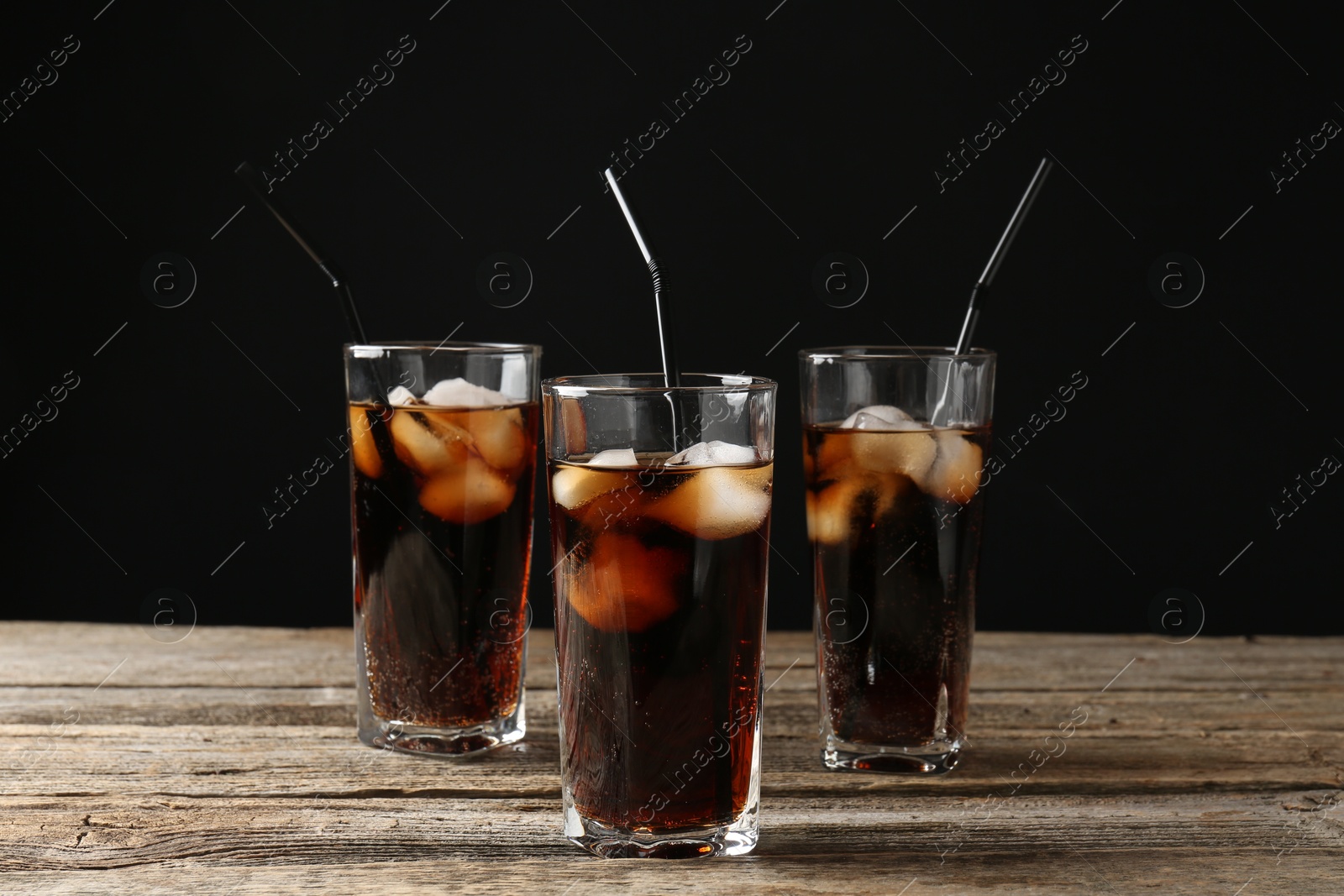 Photo of Refreshing cola with ice cubes and drinking straws in glasses on wooden table against black background