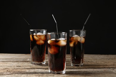 Photo of Refreshing cola with ice cubes and drinking straws in glasses on wooden table against black background