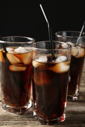 Photo of Refreshing cola with ice cubes and drinking straws in glasses on wooden table against black background, closeup