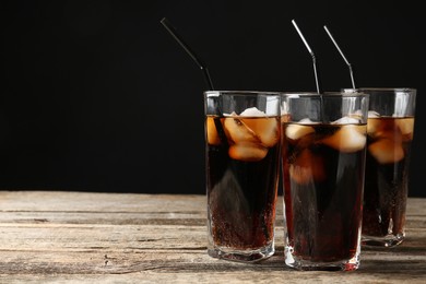 Photo of Refreshing cola with ice cubes and drinking straws in glasses on wooden table against black background, closeup. Space for text