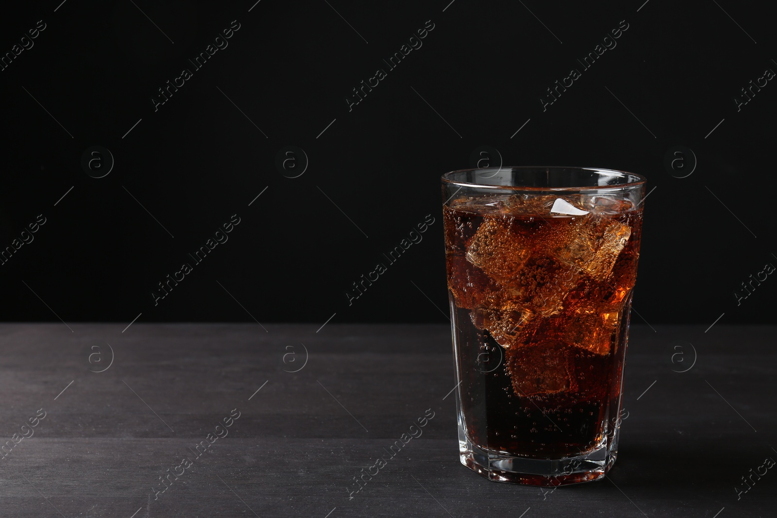 Photo of Refreshing cola with ice cubes in glass on black wooden table, closeup. Space for text