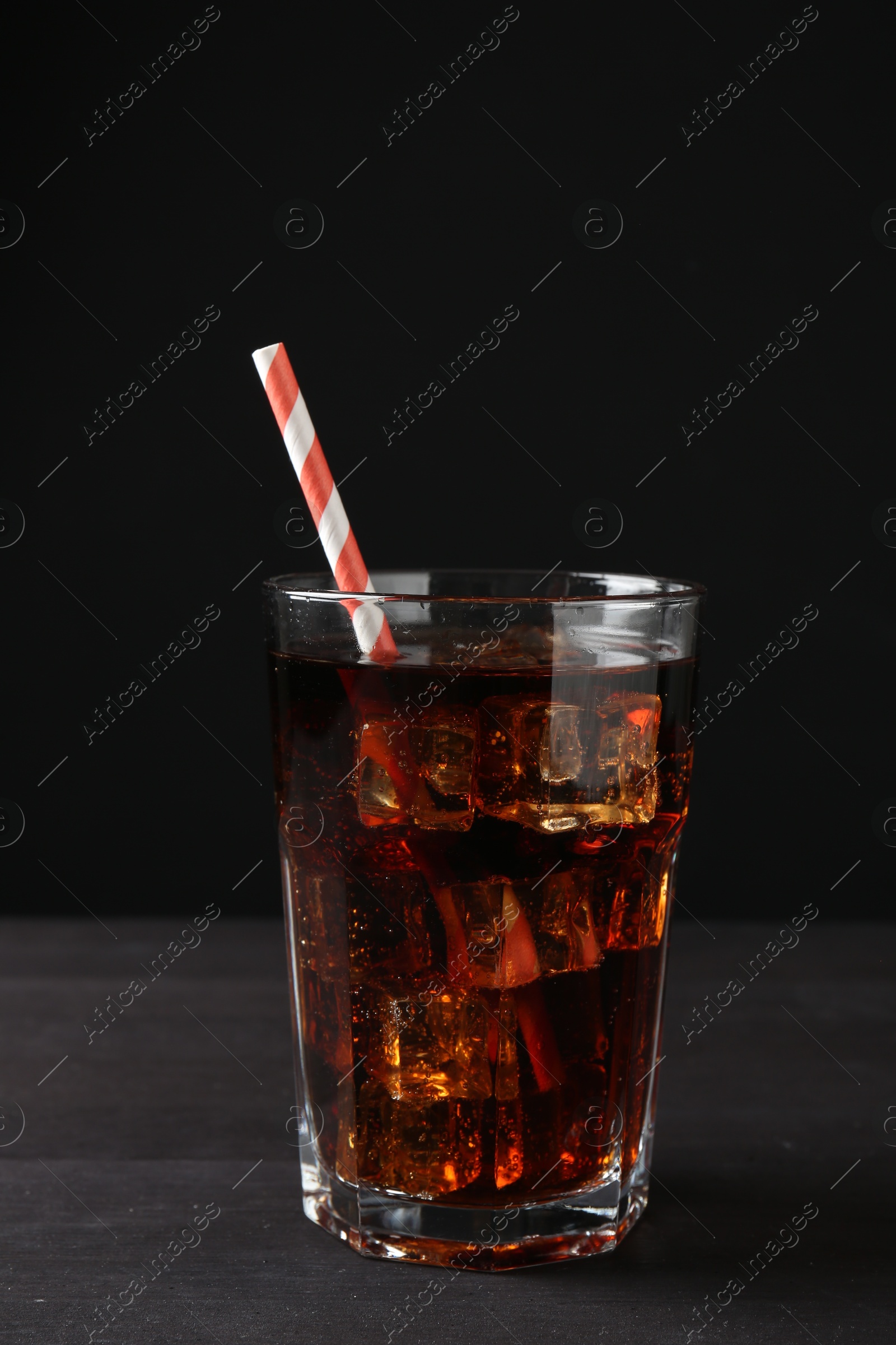 Photo of Refreshing cola with ice cubes and drinking straw in glass on black wooden table, closeup