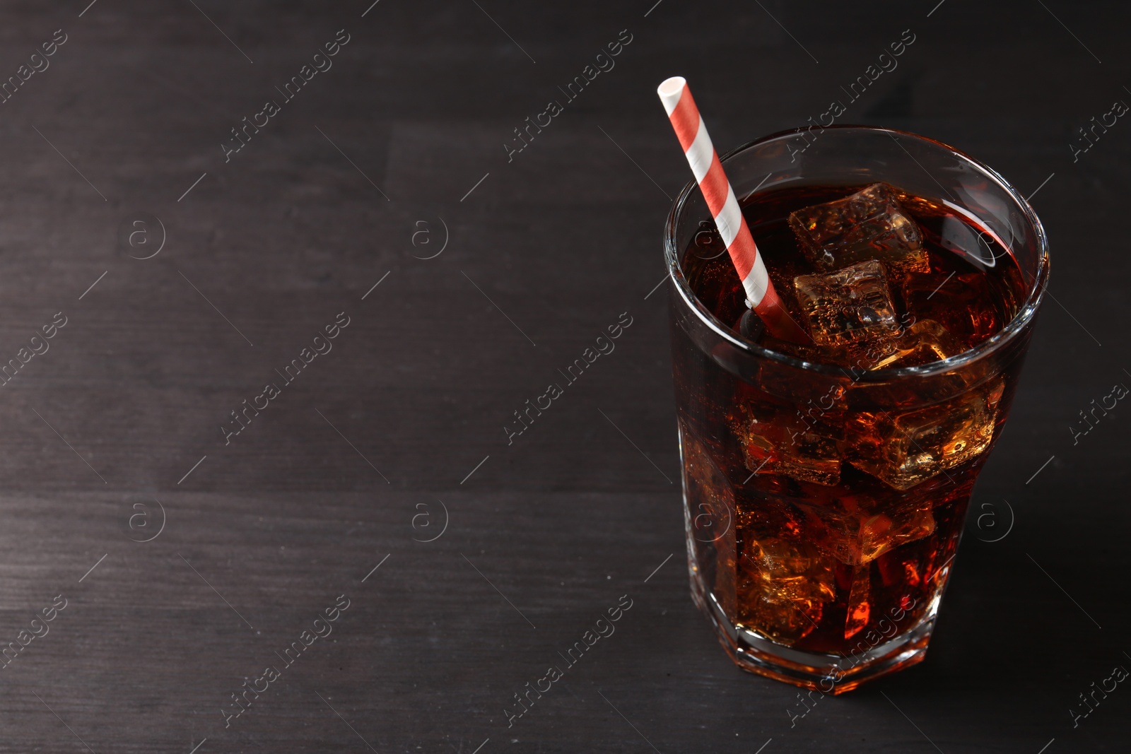 Photo of Refreshing cola with ice cubes and drinking straw in glass on black wooden table, closeup. Space for text