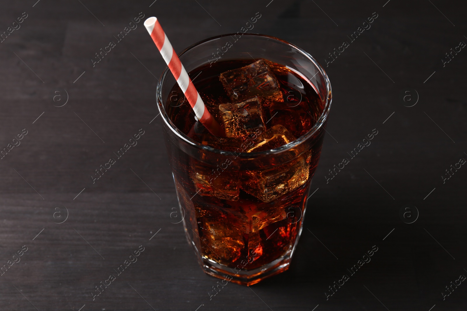 Photo of Refreshing cola with ice cubes and drinking straw in glass on black wooden table, closeup