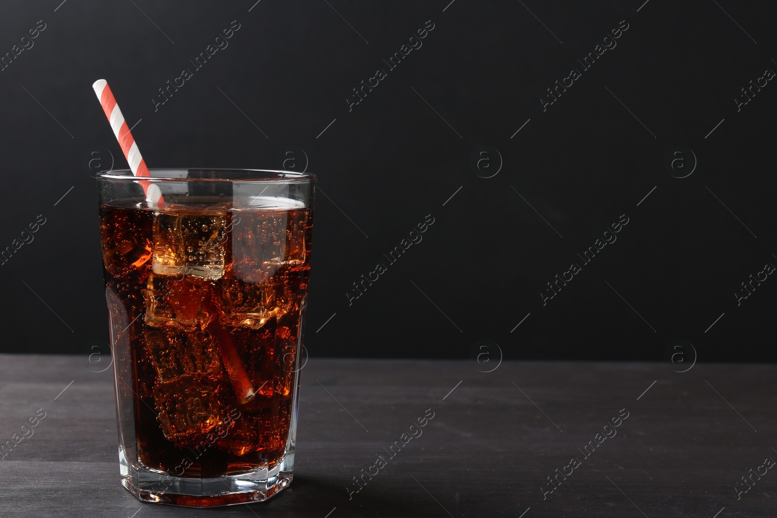 Photo of Refreshing cola with ice cubes and drinking straw in glass on black wooden table, closeup. Space for text