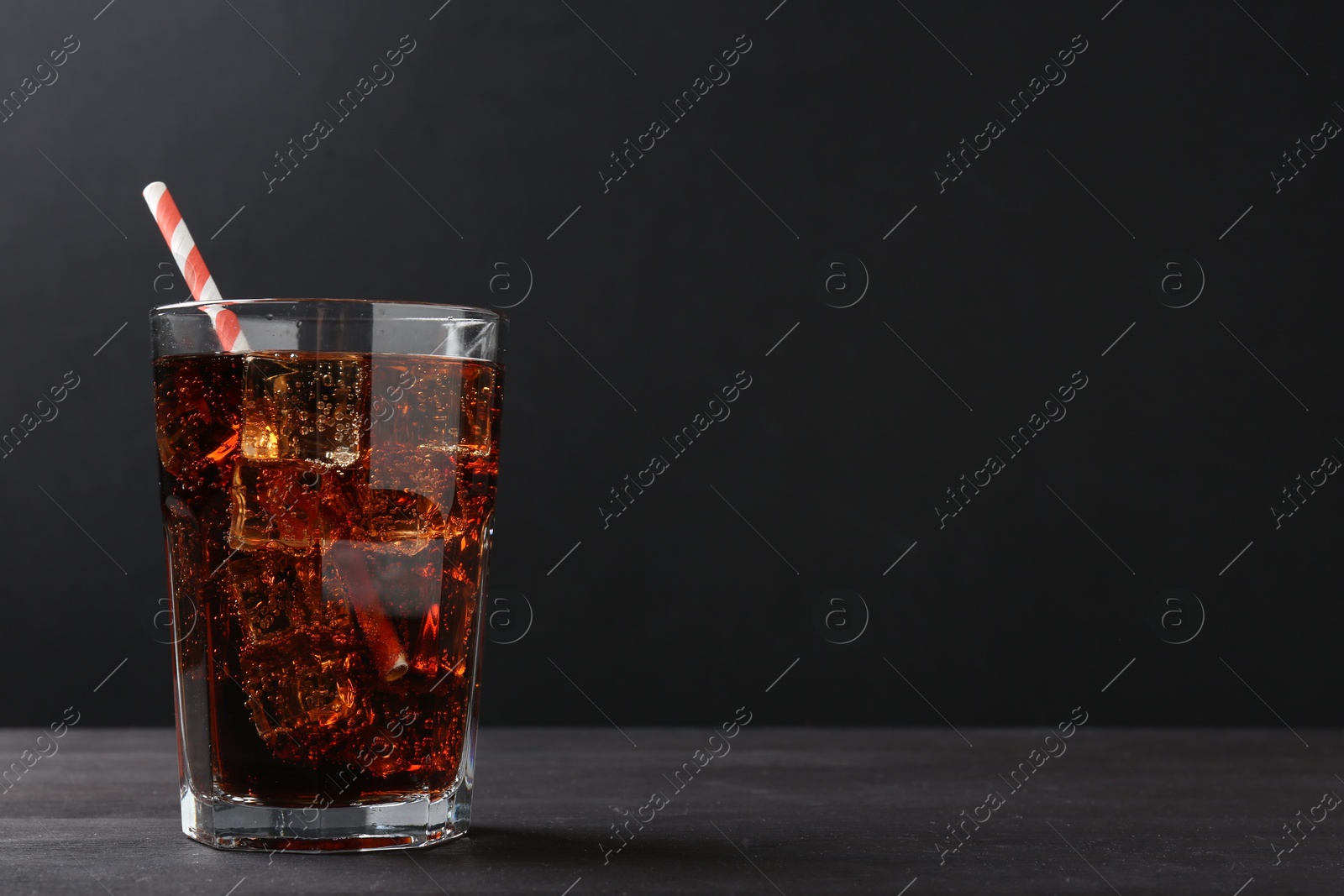 Photo of Refreshing cola with ice cubes and drinking straw in glass on black wooden table, closeup. Space for text