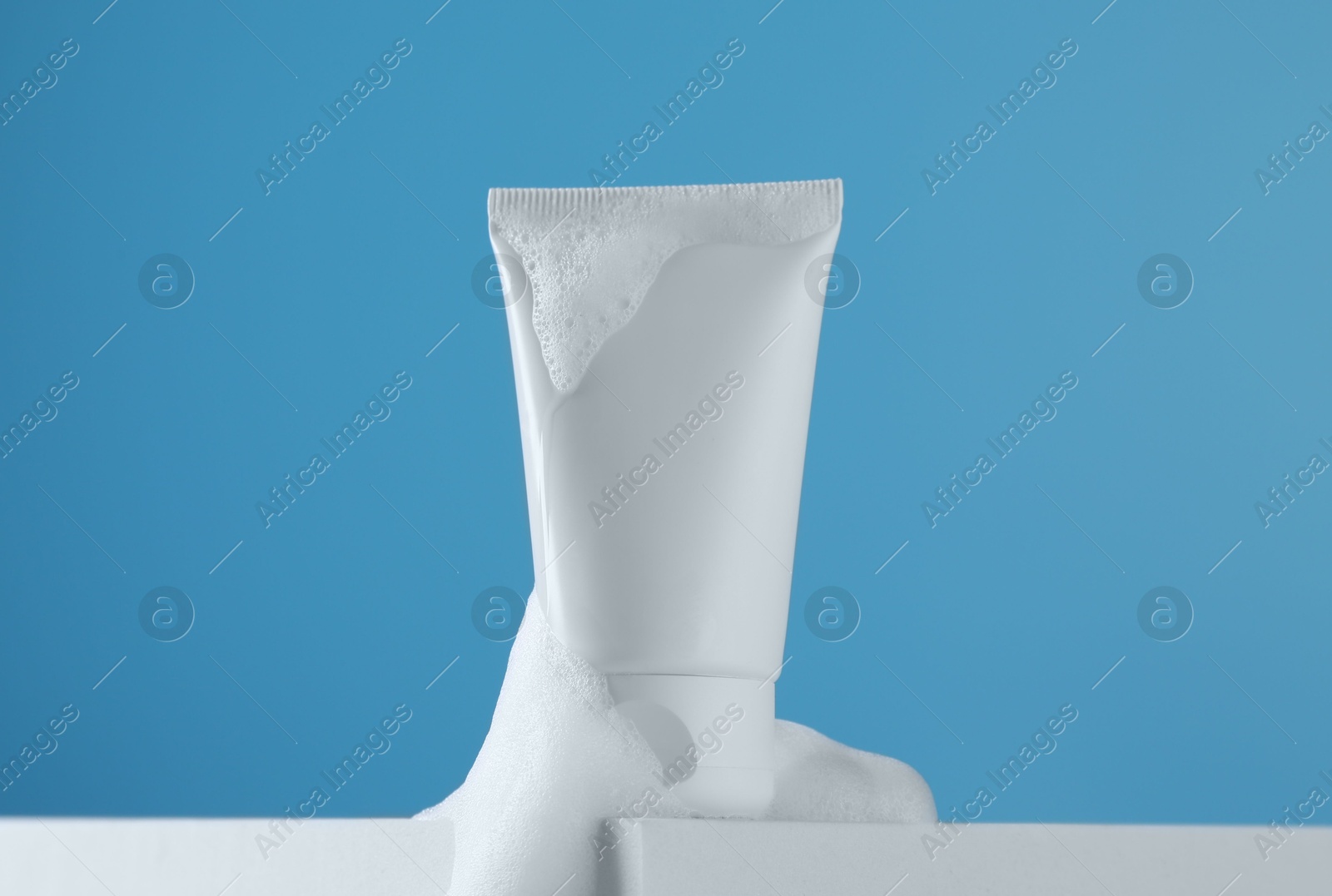 Photo of Tube of cosmetic product with foam on podium against light blue background