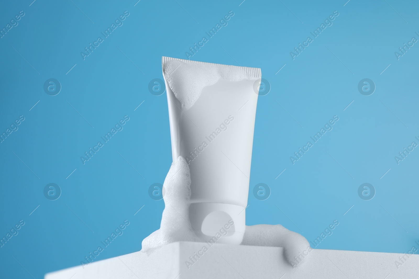 Photo of Tube of cosmetic product with foam on podium against light blue background