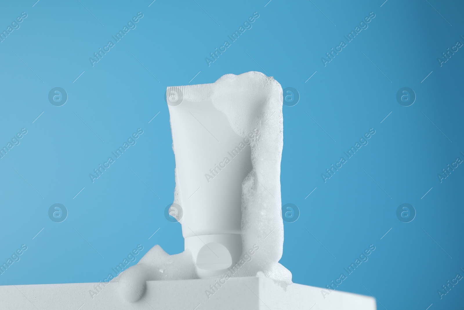 Photo of Tube of cosmetic product with foam on podium against light blue background