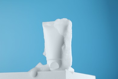 Photo of Tube of cosmetic product with foam on podium against light blue background