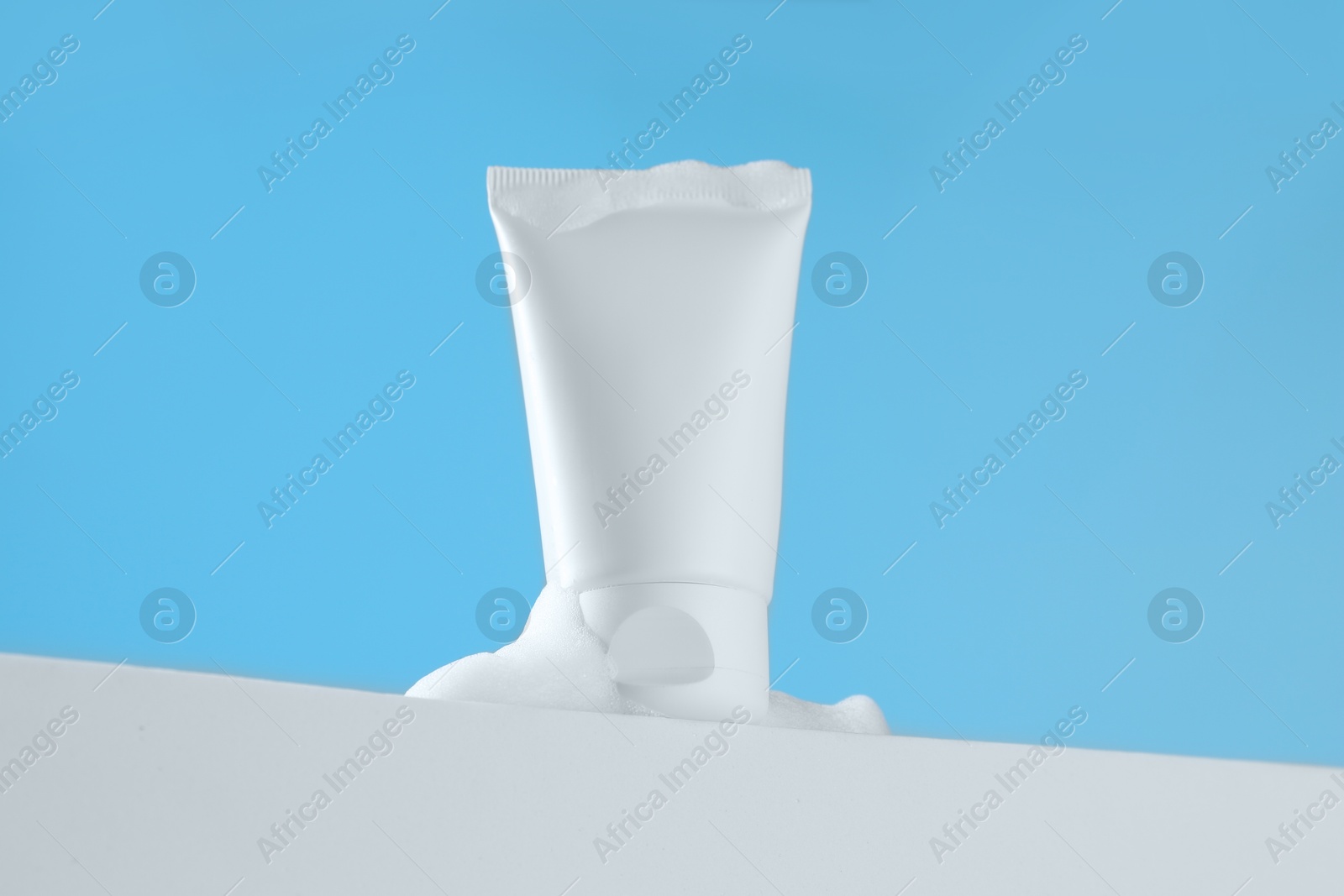 Photo of Tube of cosmetic product with foam on podium against light blue background