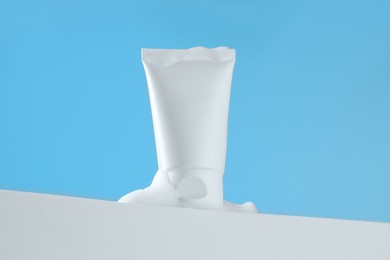 Tube of cosmetic product with foam on podium against light blue background