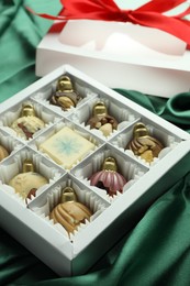 Photo of Box with tasty sweets in shape of baubles on green fabric background, closeup