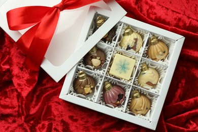 Photo of Box with tasty sweets in shape of baubles on red fabric background, top view