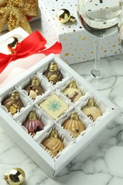 Photo of Box with tasty sweets in shape of baubles, wine and Christmas decor on white marble background, closeup