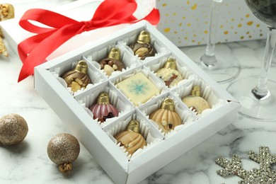 Photo of Box with tasty sweets in shape of baubles and Christmas decor on white marble background, closeup